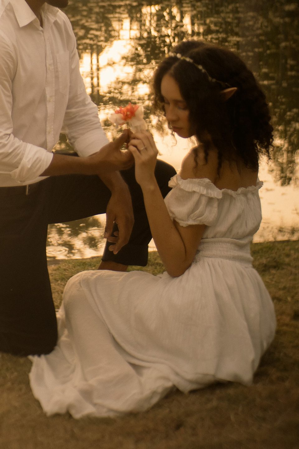Golden evening light by the water as he kneels and offers her a flower.